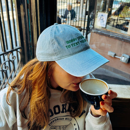 Person wearing a cap with text "Sorry I forgot to text you back", drinking from a coffee cup in an outdoor setting.