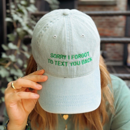 Person wearing a light gray cap with green text 'SORRY I FORGOT TO TEXT YOU BACK' outdoors.
